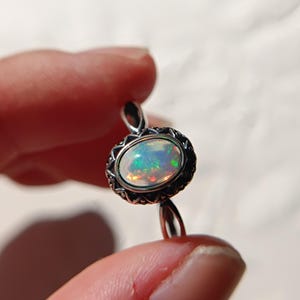 Könnte beinhalten: Ein Silberring mit einem ovalen Opal, der irisierende grüne, orangefarbene und blaue Lichtreflexe aufweist. Der Stein ist in eine verzierte, dunkel abgesetzte Silberfassung eingefasst, die ein geometrisches Muster am Rand zeigt.