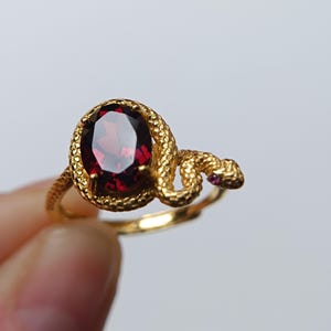 Könnte beinhalten: Ein goldener Ring mit einem strukturierten Schlangenmotiv, das sich um einen zentralen, oval geschliffenen, tiefroten Granat windet. Der Schlangenkopf ist mit einem kleinen, eingebetteten rosa Stein als Auge versehen und der Ring hat ein poliertes Goldfinish.