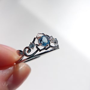 Könnte beinhalten: Ein silberfarbener Ring mit kronenartigem Design, besetzt mit einem zentralen runden blauen Edelstein und zwei kleineren klaren Steinen. Die Metallschiene weist filigrane Schnörkel und blattartige Details auf, die eine zarte, offene Struktur bilden.