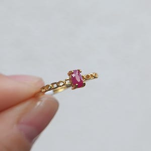 Könnte beinhalten: Ein goldener Ring mit einem kleinen, oval geschliffenen, leuchtend pink-roten Edelstein in der Mitte. Die Ringschiene hat ein filigranes, strukturiertes Kettenglied-Design, das vom zentralen Stein ausgeht und ein detailliertes Aussehen verleiht.