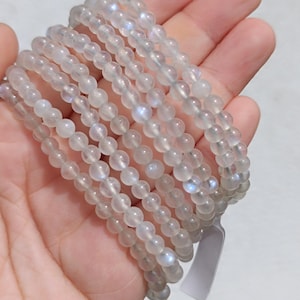 Könnte beinhalten: Ein Stapel aus mehreren Armbändern, die aus durchscheinenden, runden, hellgrauen Perlen bestehen. Jede Perle zeigt einen subtilen, irisierenden blauen Schimmer, der für Mondstein charakteristisch ist. Die Armbänder sind auf elastischem Band.