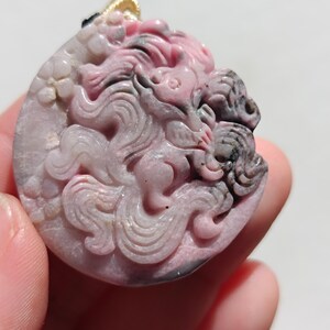 Könnte beinhalten: Ein geschnitzter Anhänger aus rosa und grauem Edelstein mit einem Relief einer mythischen Kreatur. Der Anhänger hat eine runde Form mit einer goldfarbenen Öse. Die Farbe des Steins reicht von hellrosa bis dunklerem Grau.