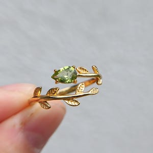 Könnte beinhalten: Ein goldfarbener Ring im Bypass-Design mit einem birnenförmigen, hellgrünen Edelstein an einem Ende und einem strukturierten Blattmotiv entlang der Ringschiene, was ein zartes botanisches Erscheinungsbild mit polierten Oberflächen erzeugt.