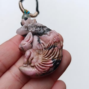 Könnte beinhalten: Ein geschnitzter Anhänger aus rosa und schwarzem Stein, der einen Hasen auf einer Mondsichel zeigt, verziert mit floralen Details. Der Anhänger hängt an einer schwarzen Kordel mit einer kleinen grünen Perle und beigem Garn am oberen Ende.