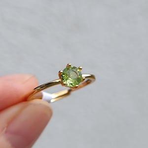 Könnte beinhalten: Ein goldfarbener Ring mit einem runden, facettierten, hellgrünen Edelstein in einer Vier-Krappen-Fassung. Die Ringschiene ist dünn und poliert, wobei ein kleines weißes Etikett am unteren Teil des Metallrings befestigt ist.