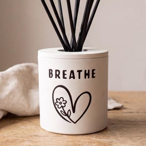 May include: A white cylindrical reed diffuser vessel featuring the black text BREATHE above a black line drawing of a heart containing a small flower. Several thin black diffuser reeds extend upwards from the centre of the container.