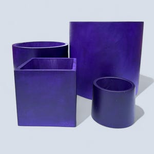 May include: Four purple containers of varying shapes and sizes. There is a tall cylinder, a square box, a short cylinder, and a small angled cylinder. The containers are all a solid, deep purple color and appear to be made of a solid material.