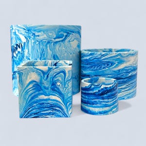 May include: A set of four blue and white marbled planters in different sizes. The planters feature a swirling pattern in shades of blue and white. The shapes include a square, a cylinder, and a short, round container.