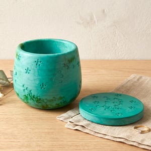 May include: A turquoise cylindrical container with a matching circular lid, both featuring a marbled texture and engraved star patterns. The container has an open top, and the lid is placed flat beside it, displaying a constellation of small stars.