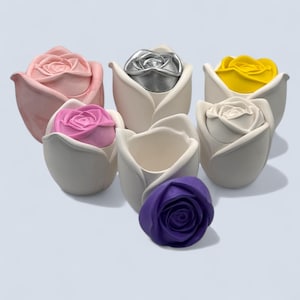 May include: A selection of ceramic rose-shaped containers in various colours. The containers are in shades of pink, white, yellow, and purple. Each rose has a lid or a centre that matches the colour of the rose.