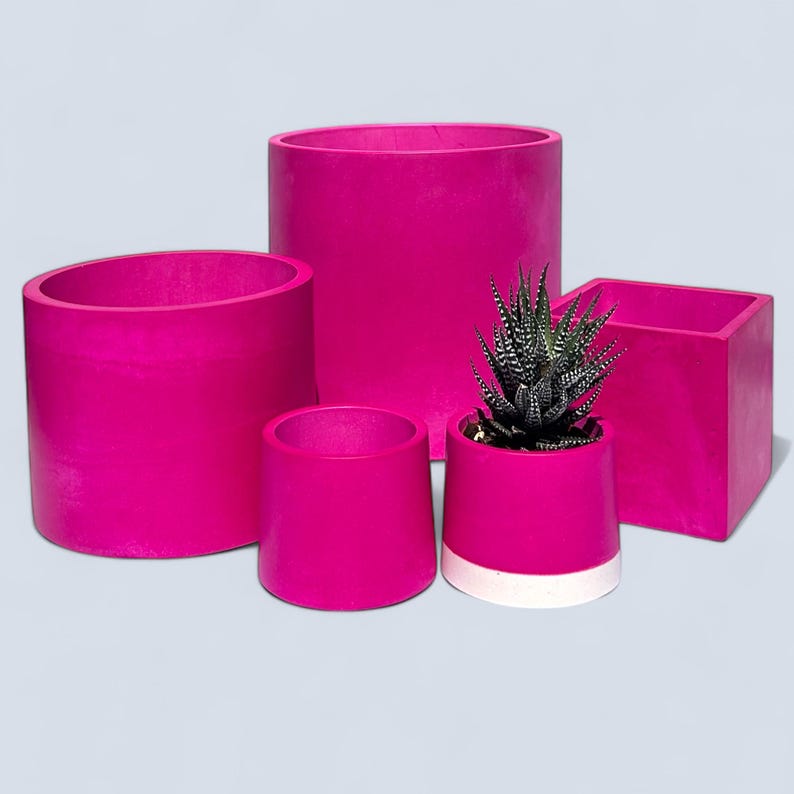 A set of 5 vibrant handmade magenta plant pots in 4 different sizes. They all have a smooth texture and a bright fuchsia colour. There is a 19cm cylindrical planter at the back, a 15cm cylindrical planter to the left, a 10cm square planter to the right and two 7.5 cm slightly tapers pots in front one with a white stripe at the bottom.