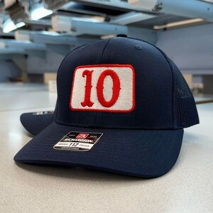 May include: Navy blue trucker hat with a white and red embroidered patch featuring the number "10". The hat has a mesh back and a curved brim. The hat is adjustable and has a tag that reads "Richardson 112 Trucker".