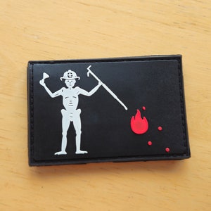 May include: Black rectangular patch featuring a white skeleton figure holding a fire axe and a pike, with a red flame and dots. The patch has a black border and is likely made of fabric or a similar material.