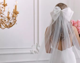 May include: A white bridal veil with a large white bow on top. The veil is made of soft tulle and has a delicate, flowing design. The bow is tied in a simple knot and adds a touch of elegance to the veil.