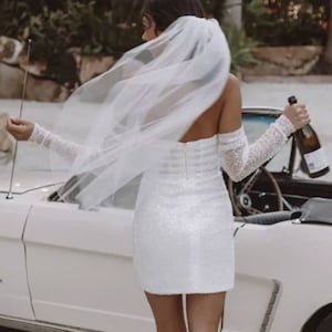 May include: A white sequined mini dress with long, sheer sleeves and a matching veil. The dress is paired with white high heels with fluffy accents. A bottle of champagne is held near a classic white convertible.