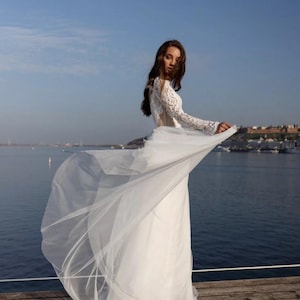 May include: A white lace wedding dress with long sleeves and a flowing tulle skirt. The dress is worn by a woman standing on a wooden jetty with a body of water in the background. The text "ToshaShop" is visible in the bottom right corner of the image.