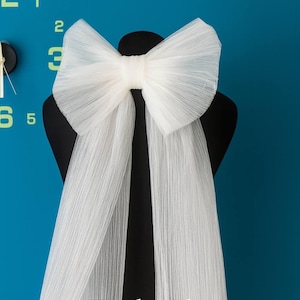 Puede incluir: Un gran lazo de tul blanco con largas cintas fluidas se muestra sobre un maniquí negro. El lazo es el punto focal, con una apariencia delicada y texturizada. El fondo es de color verde azulado sólido, con un reloj visible en la esquina superior izquierda. El texto "ToshaShop" está en la parte inferior.