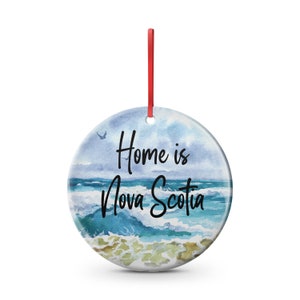 May include: Circular ceramic ornament featuring a watercolour design of ocean waves and the text "Home is Nova Scotia." The ornament has a red ribbon for hanging. The design uses shades of blue, green, and white.