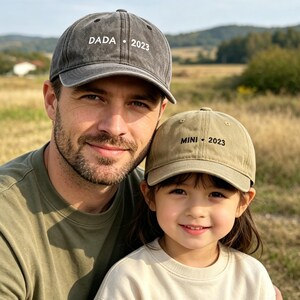 May include: A father and child are wearing matching baseball caps. The dark gray cap says "DADA 2023" and the khaki cap says "MINI 2023". They are smiling in a field on a sunny day.