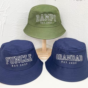 May include: Three bucket hats in the image. One is olive green with the word "BAMPI" and "EST. 2025" in white. The other two are navy blue, one with "FUNCLE" and the other with "GRANDAD", both with "EST. 2025" in white.