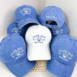 May include: A collection of baseball caps in blue and white. The blue caps feature white embroidery reading CAPE MAY EST 2026 Girls Club with a seashell icon. One blue cap shows the back with Haile's Bach text. The white cap has blue embroidery.