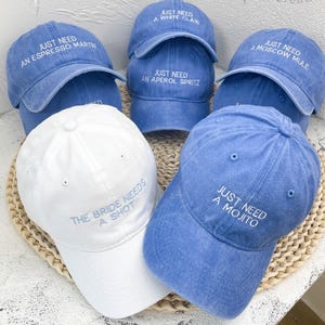 May include: A collection of six baseball caps in blue and white. Five blue caps feature white embroidered text: JUST NEED AN ESPRESSO MARTINI, JUST NEED A WHITE CLAW, JUST NEED AN APEROL SPRITZ, JUST NEED A MOSCOW MULE, and JUST NEED A MOJITO. One white cap reads THE BRIDE NEEDS A SHOT.