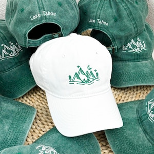 Op de afbeelding: Een verzameling groene en witte baseballpetten. De witte pet heeft een groen berg- en boomontwerp met een maan en sterren. De groene petten hebben de tekst "Lake Tahoe" en een bergontwerp.