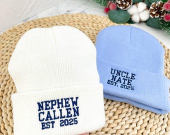 Può includere: Due berretti di lana, uno bianco e uno azzurro chiaro, con testo ricamato. Il berretto bianco dice "Nephew Callen Est. 2025" e il berretto azzurro dice "Uncle Nate Est. 2025".