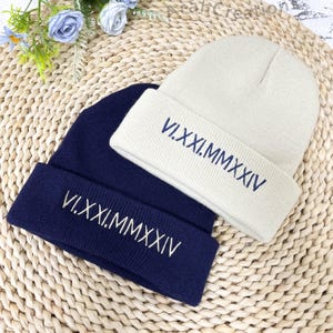 May include: Two knitted beanies, one navy blue and one beige, with the Roman numeral inscription "VI.XXI.MMXXIV" embroidered on each.