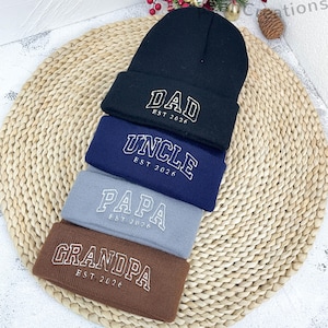 Peut inclure: Une pile de quatre bonnets et bandeaux en noir, bleu marine, gris et marron. Chaque article porte une broderie blanche : "DAD", "UNCLE", "PAPA" et "GRANDPA", avec "EST 2026" en dessous. Ils sont présentés sur un tapis tressé.