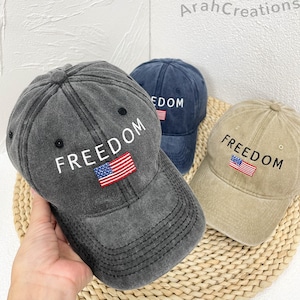 Può includere: Tre cappellini da baseball grigi, blu navy e beige. Ogni cappellino presenta la parola "FREEDOM" ricamata in bianco, insieme a una piccola toppa con la bandiera americana. I cappellini sono realizzati in un tessuto usurato e strutturato.