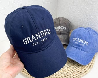May include: Three baseball caps with embroidered text. The first cap is navy blue with white text that reads "GRANDAD EST. 2024". The second cap is gray with white text that reads "GRANDPA EST. 2023". The third cap is light blue with white text that reads "GRANDPA EST. 2025".