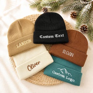 Könnte beinhalten: Fünf Strickmützen in Schwarz, Hellbraun, Creme, Braun und Blaugrün. Die schwarze Mütze trägt den Text Custom Text, die hellbraune LANDON, die cremefarbene Oliver, die braune SLOAN und die blaugrüne zeigt ein Berggrafik-Logo mit dem Text Custom Logo.
