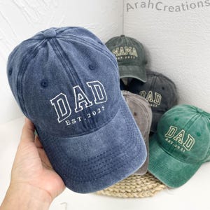 May include: A set of four denim baseball caps with the word "DAD" embroidered on the front in white. Each cap has a different colour and a different year embroidered on the back. The caps are arranged in a circle on a woven surface.