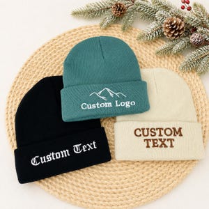 Puede incluir: Tres gorros de punto en negro, verde azulado y crema. El gorro negro presenta texto bordado en blanco que dice Custom Text. El gorro verde azulado muestra un gráfico de montaña blanco y el texto Custom Logo. El gorro crema tiene texto bordado en marrón que dice CUSTOM TEXT.