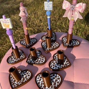 May include: A collection of pens and pen holders. The pens feature pink or blue rhinestone barrels, decorative beads, and toppers including bows or signs reading "I'd rather be Thrifting". The brown heart-shaped holders have a white leopard print base.