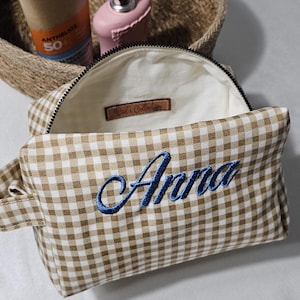 May include: A tan and white gingham cosmetic bag with the name "Anna" embroidered in blue. The bag is open, revealing a light interior. A tube of sunscreen and a pink perfume bottle are in a woven basket in the background.