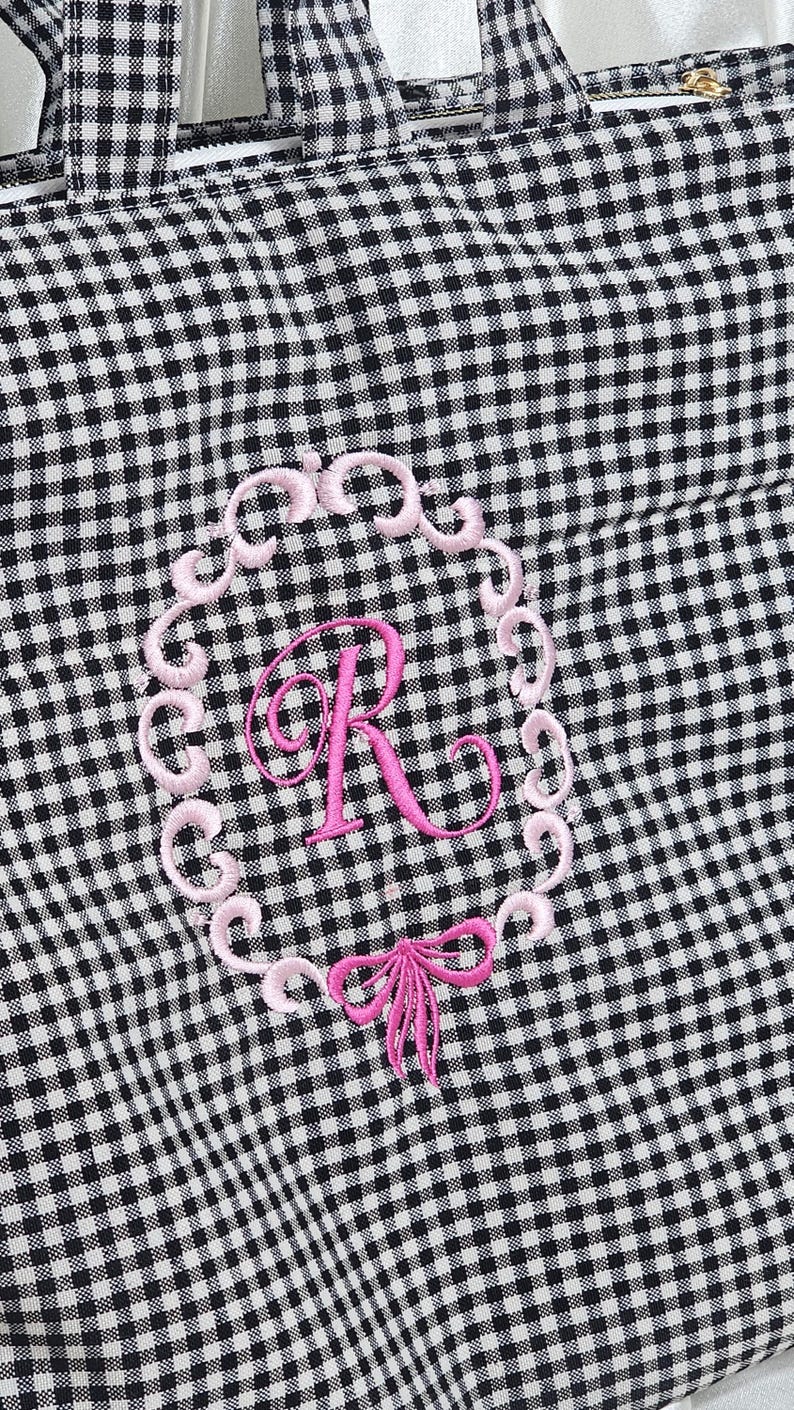 May include: Black and white gingham tote bag with a pink embroidered initial "R" inside a decorative frame. A pink bow is embroidered below the initial. The bag has black handles and a zipper closure.
