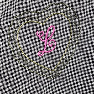 May include: Black and white gingham tote bag with a heart-shaped embroidered patch. The patch features a pink letter "L" inside a gold outline, surrounded by a scalloped edge. The bag's handles are visible at the top.