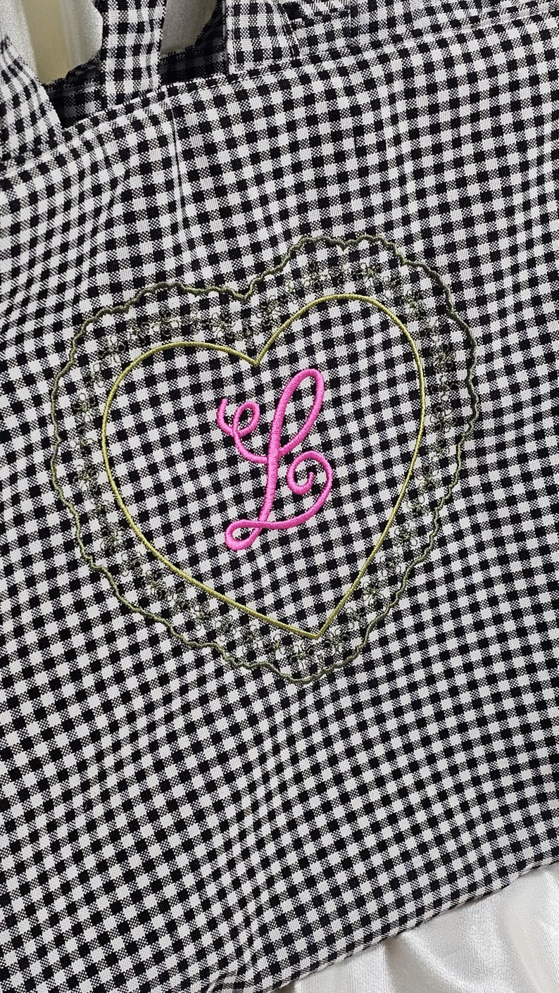 May include: Black and white gingham tote bag with a heart-shaped embroidered patch. The patch features a pink letter "L" inside a gold outline, surrounded by a scalloped edge. The bag's handles are visible at the top.