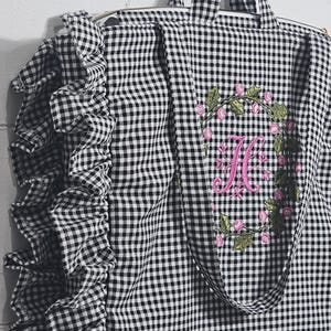 May include: Black and white gingham tote bag with a ruffle detail on the side. The bag features a pink floral wreath with a pink embroidered letter "H". The tote has two handles and a zipper closure.