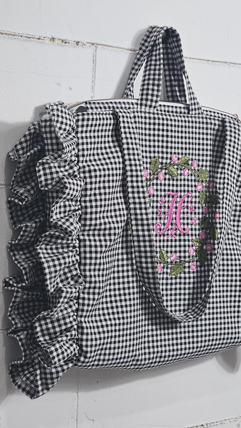May include: Black and white gingham tote bag with a ruffle detail on the side. The bag features a pink floral wreath with a pink embroidered letter "H". The tote has two handles and a zipper closure.