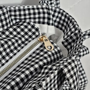 May include: Close-up of a black and white gingham tote bag. The bag features a gold zipper and a white interior. The bag has a classic checkered pattern and is a stylish accessory.