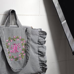 May include: Black and white gingham tote bag with a floral wreath and the letter "H" embroidered in pink. The bag has a ruffled edge and sturdy handles.