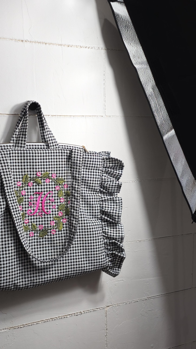 May include: Black and white gingham tote bag with a floral wreath and the letter "H" embroidered in pink. The bag has a ruffled edge and sturdy handles.