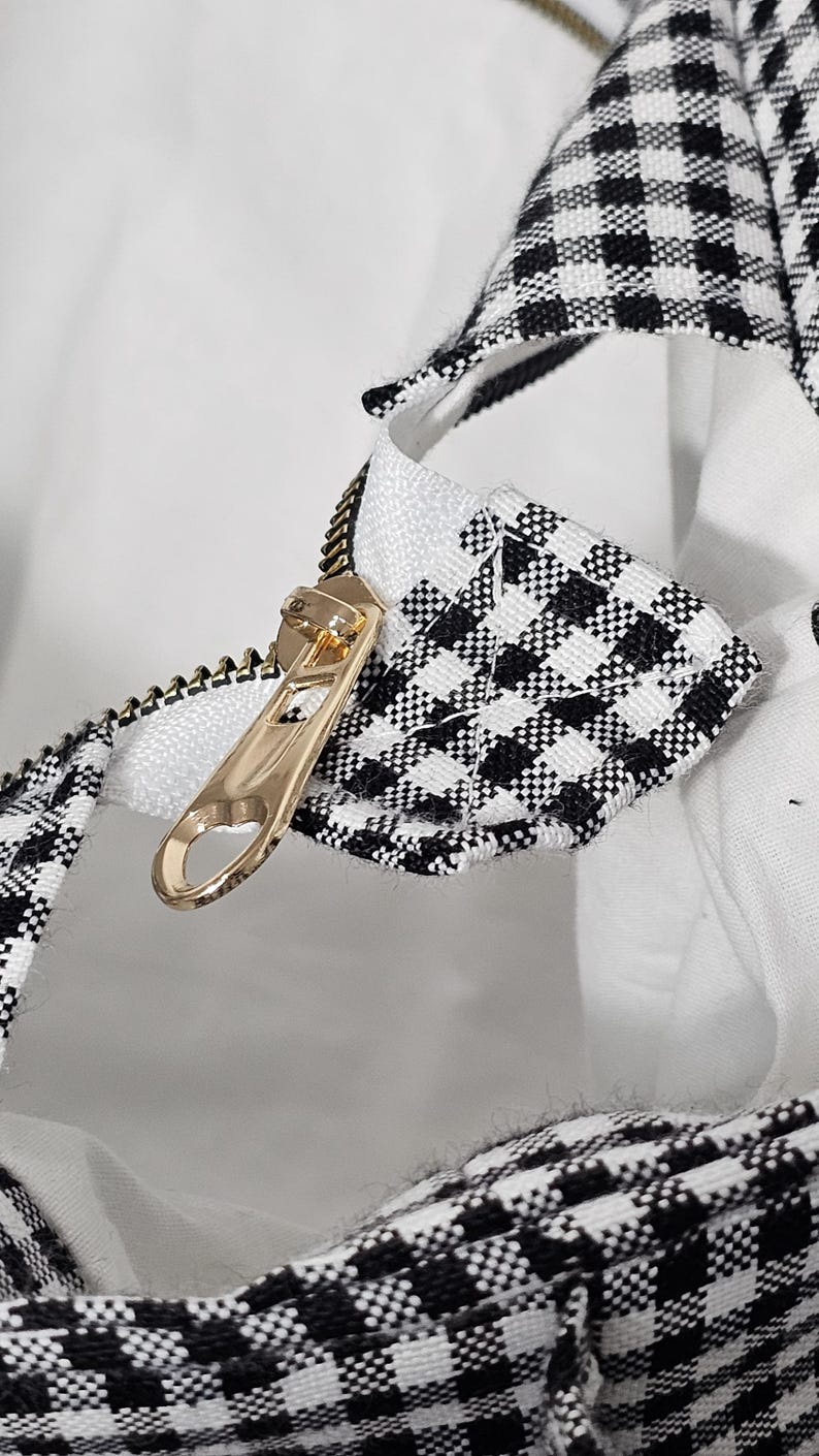 May include: Close-up of a gold zipper on a white and black checkered fabric. The zipper is partially open, revealing a white interior. The fabric has a classic gingham pattern.