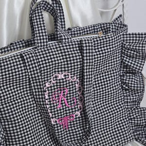 May include: A black and white gingham tote bag with a pink embroidered initial "R" in a decorative frame. The bag has a zipper closure, ruffled edges, and matching handles. The background is a white satin fabric.