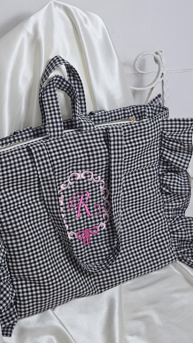 May include: A black and white gingham tote bag with a pink embroidered initial "R" in a decorative frame. The bag has a zipper closure, ruffled edges, and matching handles. The background is a white satin fabric.