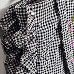 May include: Black and white gingham fabric with a ruffled edge. The fabric is part of a garment, possibly a top or dress, with a floral embroidery detail in pink and green.