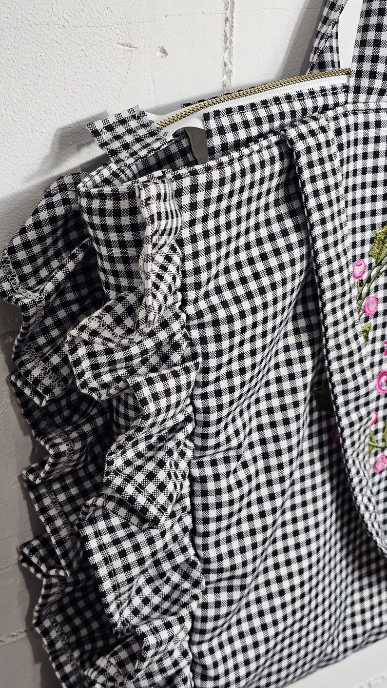 May include: Black and white gingham fabric with a ruffled edge. The fabric is part of a garment, possibly a top or dress, with a floral embroidery detail in pink and green.